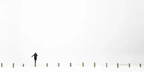 Osoba w czarnym płaszczu balansuje na słupku w śnieżnym krajobrazie. Minimalistyczna, high key fotografia.