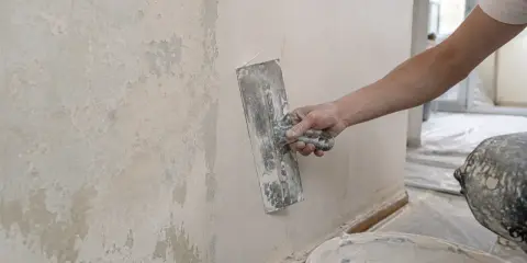 Ręka trzymająca kielnię wygładza tynk na ścianie. Widać starą, łuszczącą się farbę obok.