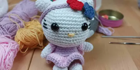Muñeca de Hello Kitty tejida a crochet, con vestido lila y lazo rojo y azul. Hilos y agujas de crochet alrededor, perfectos para crear tus propios hello kitty crochet patrones español.