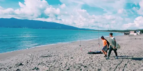 Due ragazzi passeggiano sulla spiaggia della Maremma Toscana, uno con una tavola da surf, mentre il mare azzurro lambisce la riva.