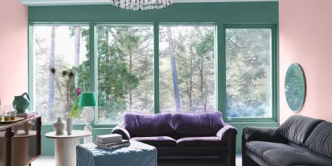 Ett vardagsrum med art deco inredning, mjuklila soffa, mörkgrå soffa, turkos pall och en stor fönsterutsikt över skogen.