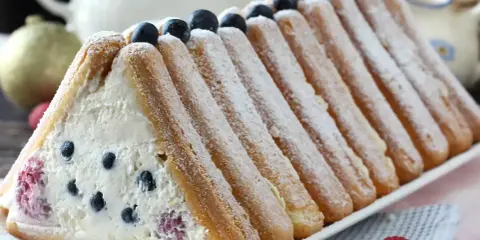 Délicieuse recette buche fruits rouges : un gâteau léger garni de crème et de baies, entouré de biscuits à la cuillère saupoudrés de sucre glace.