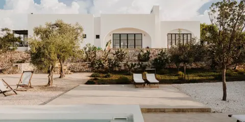Piscina e lettini in una delle masserie famose puglia, con ulivi e cactus sotto un cielo azzurro con nuvole bianche.