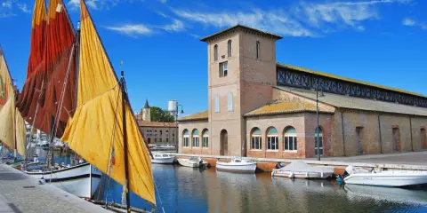 Vele colorate di barche tradizionali ormeggiate nel porto canale di Cervia mare, con l'antico edificio delle saline sullo sfondo.