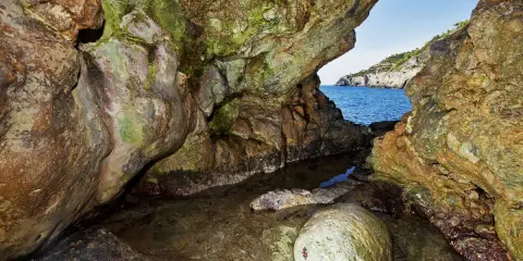 L'entrata di una grotta marina, la cala del bove, con rocce muschiose e un piccolo specchio d'acqua che riflette il blu del mare e il cielo.