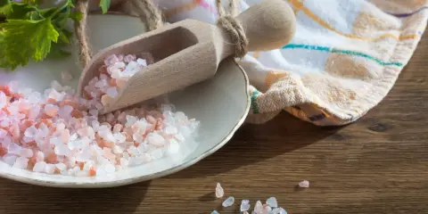 Różowa sól kamienna w drewnianej łyżce na talerzu, obok zielona gałązka i ściereczka.