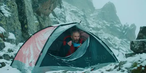 Mężczyzna w namiocie, jedzący posiłek, w zimowej scenerii górskiej. Scena jak z filmów wspinaczkowych.