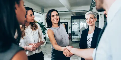 Uśmiechnięte kobiety w biurze, jedna z nich ściska dłoń partnerowi, symbolizując udaną współpracę w spółce partnerskiej.
