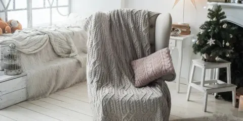 Manta de punto gris con trenzado, estilo plaid Bassetti, sobre sillón junto a árbol de Navidad.