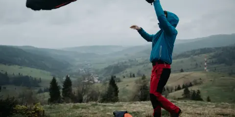 Osoba w niebieskiej kurtce i czerwonych spodniach rozkłada śpiwór puchowy czy syntetyczny na tle górskiego krajobrazu.