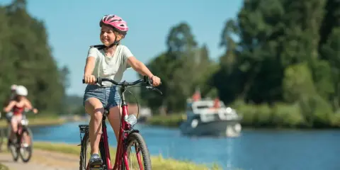 Glad flicka på cykel med hjälm njuter av en cykelsemester med barn längs en kanal. En båt syns i bakgrunden.