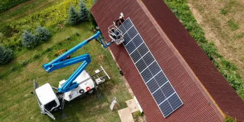 Installatörer monterar solpaneler på ett tak med hjälp av en kranbil. Solpaneler omvandlar solljus till likström (DC), som sedan kan omvandlas till växelström (AC) för användning i hemmet.