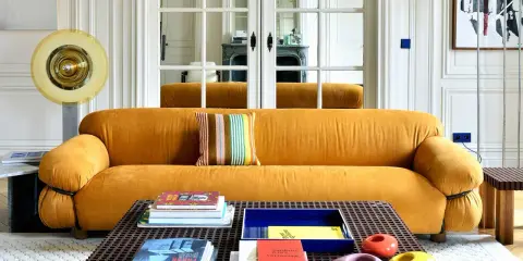 Divano senape con cuscino a righe colorate. Tavolino con libri e oggetti decorativi. Abbinamenti moderni e audaci.