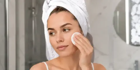 Kobieta z ręcznikiem na głowie delikatnie zmywa twarz wacikiem. Dowiedz się, jak zmyć samoopalacz, by uzyskać naturalny efekt.