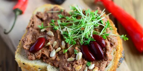 Pyszna pasta z czerwonej fasoli na chlebie, ozdobiona kiełkami i nasionami. Obok leżą czerwone papryczki chili.
