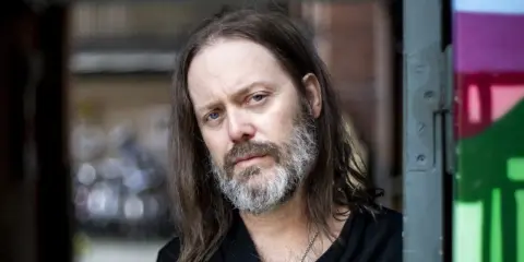 Niclas Frisk med långt hår och skägg, blickar mot kameran.