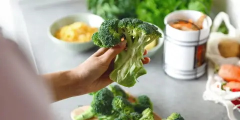Mano sosteniendo un brócoli fresco, listo para preparar deliciosas recetas de brócoli. En el fondo, queso rallado y zanahorias.