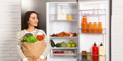 Kobieta z torbą zakupów obok otwartej lodówki pełnej jedzenia. Zastanawiasz się, co postawić na lodówce w kuchni? Może świeże warzywa i owoce?