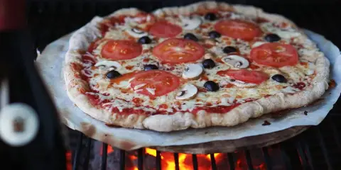 En hemmagjord pizza med tomater, champinjoner och oliver grillas över glödande kol. Perfekt för ett italiensk pizzadeg recept.