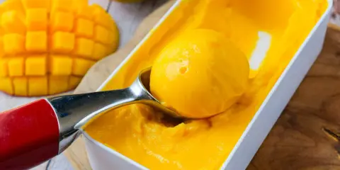 Une cuillère prélève une boule de sorbet mangue crémeux d'une boîte. Des morceaux de mangue fraîche sont visibles à côté.