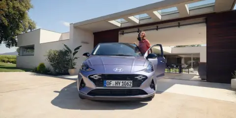 Mujer sonriente junto a un coche alto y morado, aparcado frente a una casa moderna con puertas abiertas.