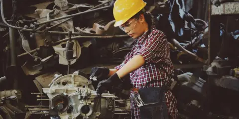 Mujer con casco amarillo y gafas de seguridad trabajando en un motor. Usa guantes negros y un delantal de mezclilla, demostrando el uso de los equipos de protección individual.