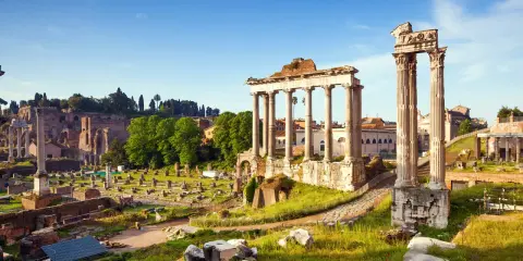 Antiche rovine del Foro Romano illuminate dal sole, con colonne imponenti e resti di templi sotto un cielo azzurro.