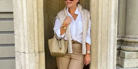 Elegancka kobieta w stylu moda 50 plus, w beżowych spodniach i białej koszuli, z torebką i swetrem narzuconym na ramiona.
