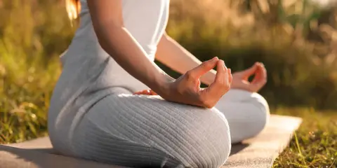 En person sitter i lotusställning på en matta utomhus, med händerna i meditation. Solen lyser genom gräset och skapar en varm atmosfär för morgonmeditation.