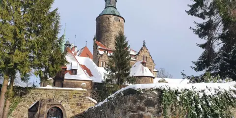 Zamek Czocha zimową porą, otoczony śniegiem i iglastymi drzewami. Kamienny mostek prowadzi do bramy.
