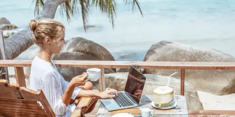 Kvinna med kaffe och laptop på en strandterrass, drömmen om ett digitalt nomad jobb.