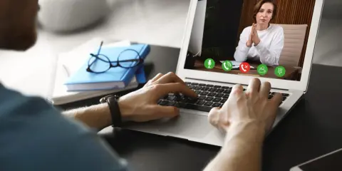 Mężczyzna prowadzi wirtualną superwizję przez laptopa. Na ekranie widać kobietę z dłońmi złożonymi w geście modlitwy.