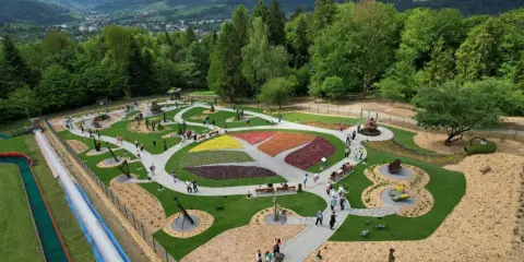 Widok z góry na park z kolorowymi rabatami kwiatowymi i rzeźbami. Widać ścieżki, ławki i ludzi spacerujących po terenie. W tle góry i las.