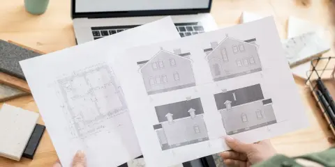 Rysunki domów i rzut parteru, przygotowane do uzyskania pozwolenia na budowę. Na biurku laptop i próbki materiałów.
