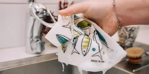 En hand håller en disktrasa i linne med fiskmönster under rinnande vatten i ett kök.