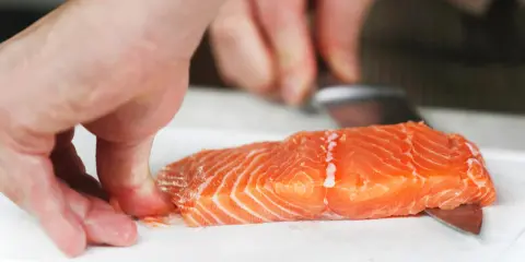 Hände halten ein Lachsfilet, während ein Messer die Haut vom Lachs entfernt.
