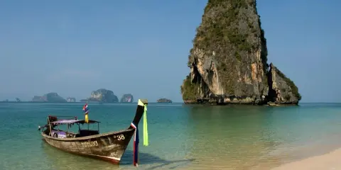 En traditionell longtailbåt guppar i det turkosa vattnet vid en strand i Thailand. I bakgrunden reser sig en grönskande klippö.