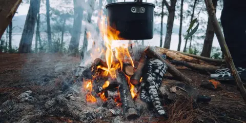 Początek przygody z gotowaniem na ogniu. Garnek wisi nad płomieniami, a w tle las i jezioro.