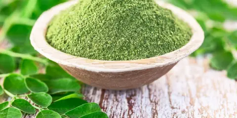 Moringa w proszku w drewnianej misce, otoczona świeżymi liśćmi. Zanim zaczniesz stosować, sprawdź moringa przeciwwskazania.