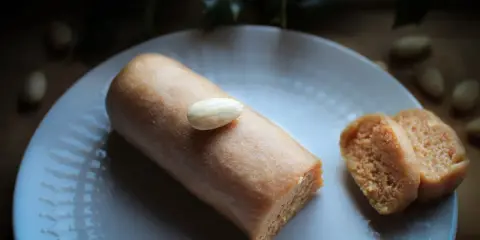 Un bâtonnet de pâte d'amande, garni d'une amande entière, repose sur une assiette blanche. À côté, deux tranches de ce délice, parfait pour un fond de tarte amande.
