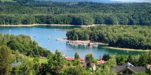 Malowniczy widok na jezioro z żaglówkami, otoczone lasami. W oddali widać góry, a na pierwszym planie domki letniskowe. Idealne miejsce na wypoczynek nad wodą, przypominające solina plaża.