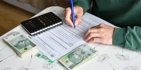 Osoba wypełnia dokumenty dotyczące umowy zlecenia i ubezpieczenia, obok leżą pieniądze i kalkulator.