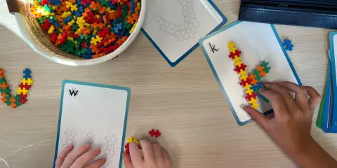 Kinderen bouwen met kleurrijke stukjes aan patronen op kaarten. Een mand vol bouwstenen ligt klaar voor meer taal spelletjes.