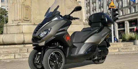 Trójkołowy skuter Peugeot Metropolis 400 na tle zabytkowej kamienicy i pomnika.