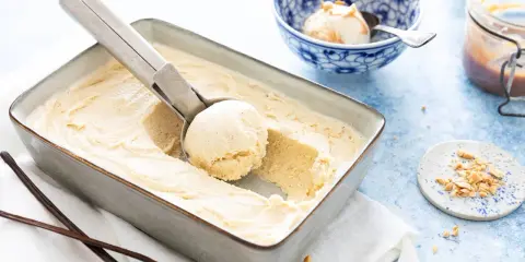 Une cuillère prélève une boule d'une glace vanille maison crémeuse dans un plat. Des gousses de vanille et des éclats de cacahuètes sont à proximité.