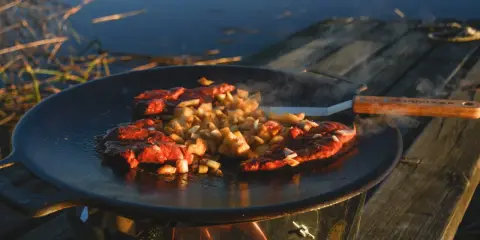 Kött och potatis steks på en rostig muurikka vid vattnet. En stekspade med trähandtag ligger bredvid.