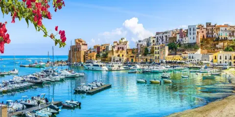 Un pittoresco porto con barche ormeggiate, case colorate su una collina e fiori rossi in primo piano, un'immagine che evoca la bellezza di Sardegna e Sicilia.