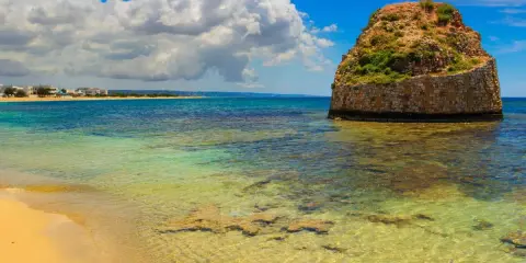 L'isola della fanciulla, una torre rotonda in pietra nel mare cristallino, con una spiaggia dorata e case in lontananza sotto un cielo azzurro con nuvole bianche.