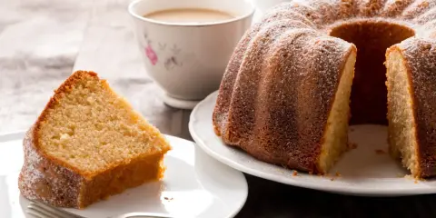Una fetta di torta al latte soffice e dorata, cosparsa di zucchero a velo, accanto a una ciambella intera e una tazza di tè.