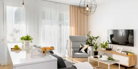 Jasny salon z białymi ścianami, szarą sofą, fotelem i telewizorem. Na blacie wyspy kuchennej owoce i pieczywo.